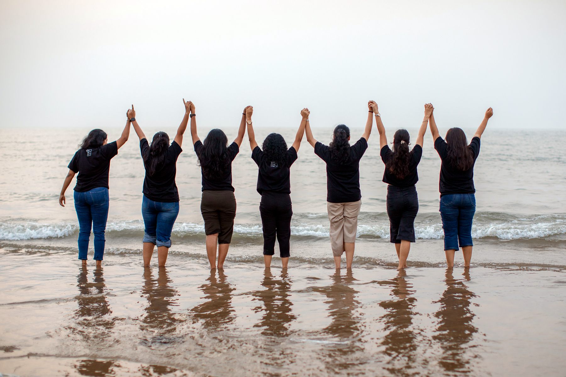 Seven women at the shoreline, hands joined and raised, facing the sea